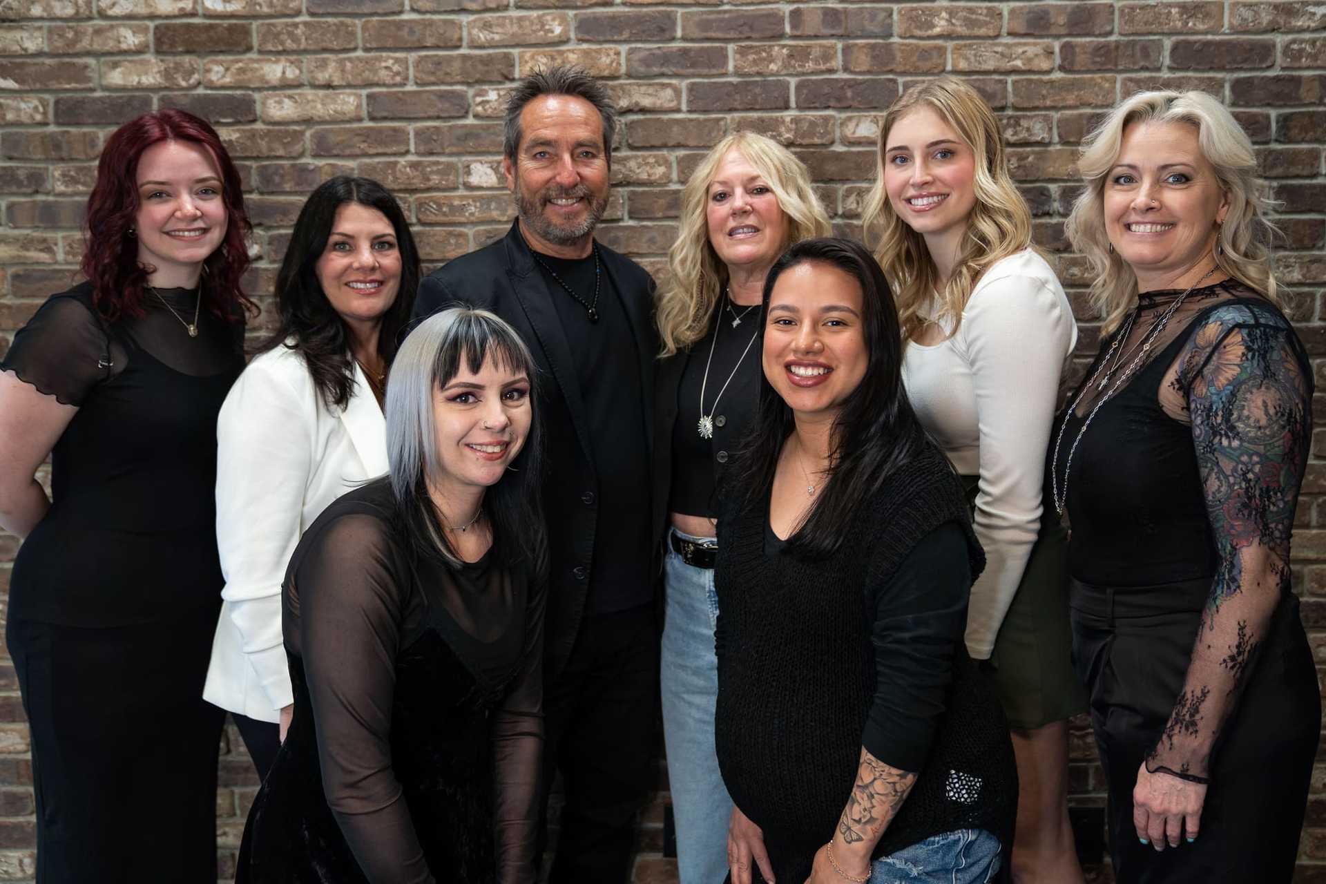 Group of eight smiling people posing in front of a brick wall.