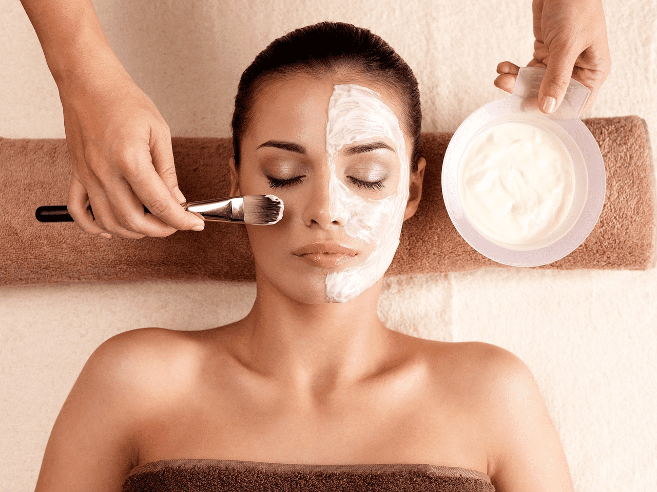 Facial treatment with brush and cream, half face applied, person relaxing on a spa bed.